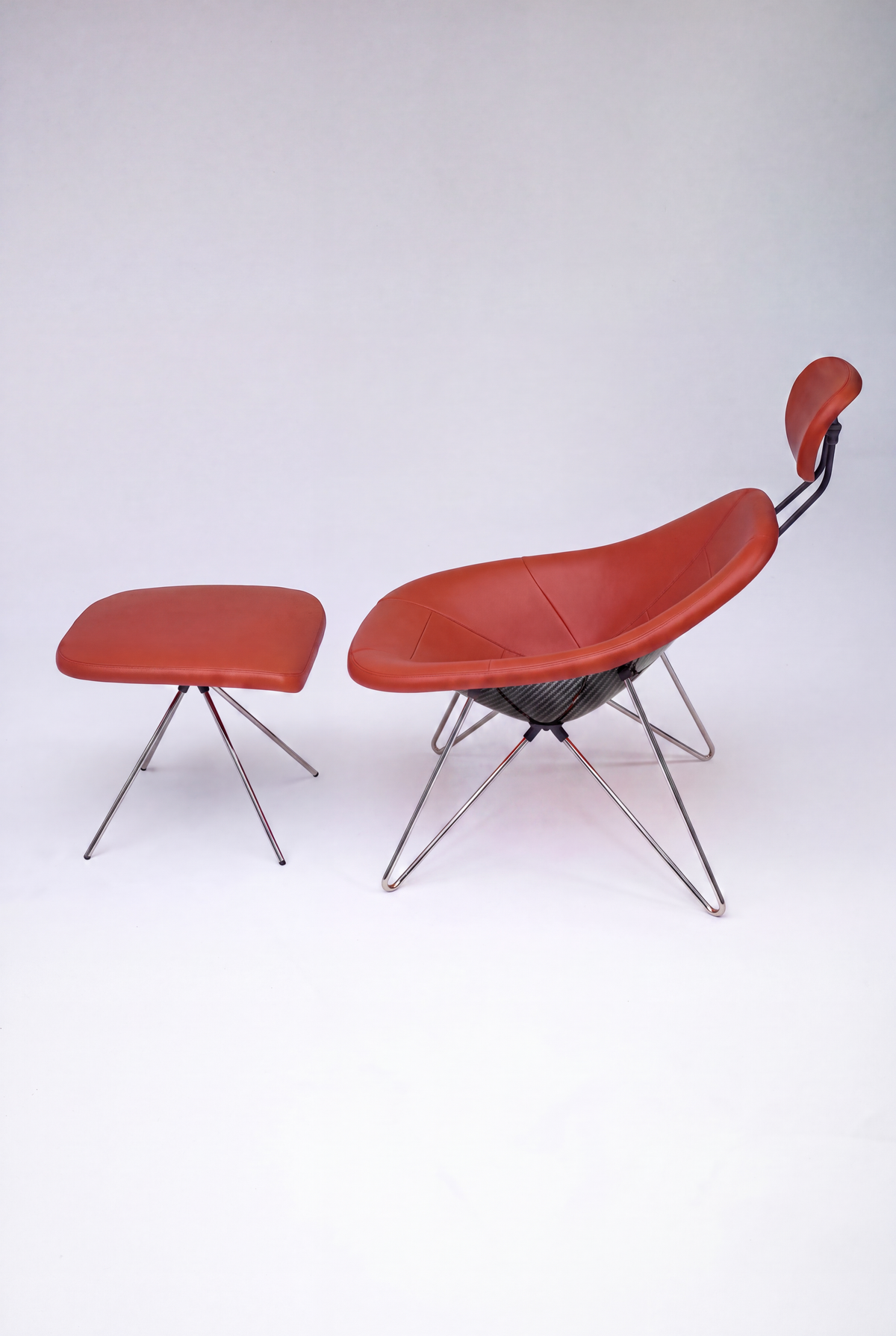 House Dé Stains bespoke automotive furniture: Full set view of the Maniac Lounge Chair and matching ottoman featuring burnt-orange leather upholstery, a hand-finished carbon fiber composite shell, and polished chrome tubular steel frames.