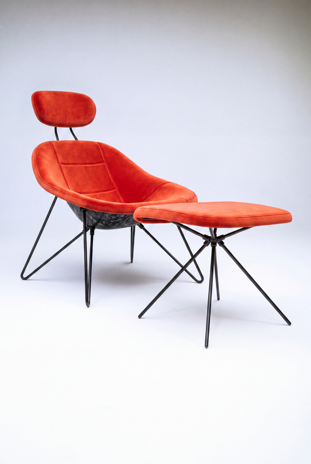 House Dé Stains bespoke automotive furniture: Three-quarter view of the Maniac Lounge Chair and matching ottoman featuring bright orange-red velvet upholstery, a hand-finished carbon fiber composite shell, and matte black tubular steel frames.