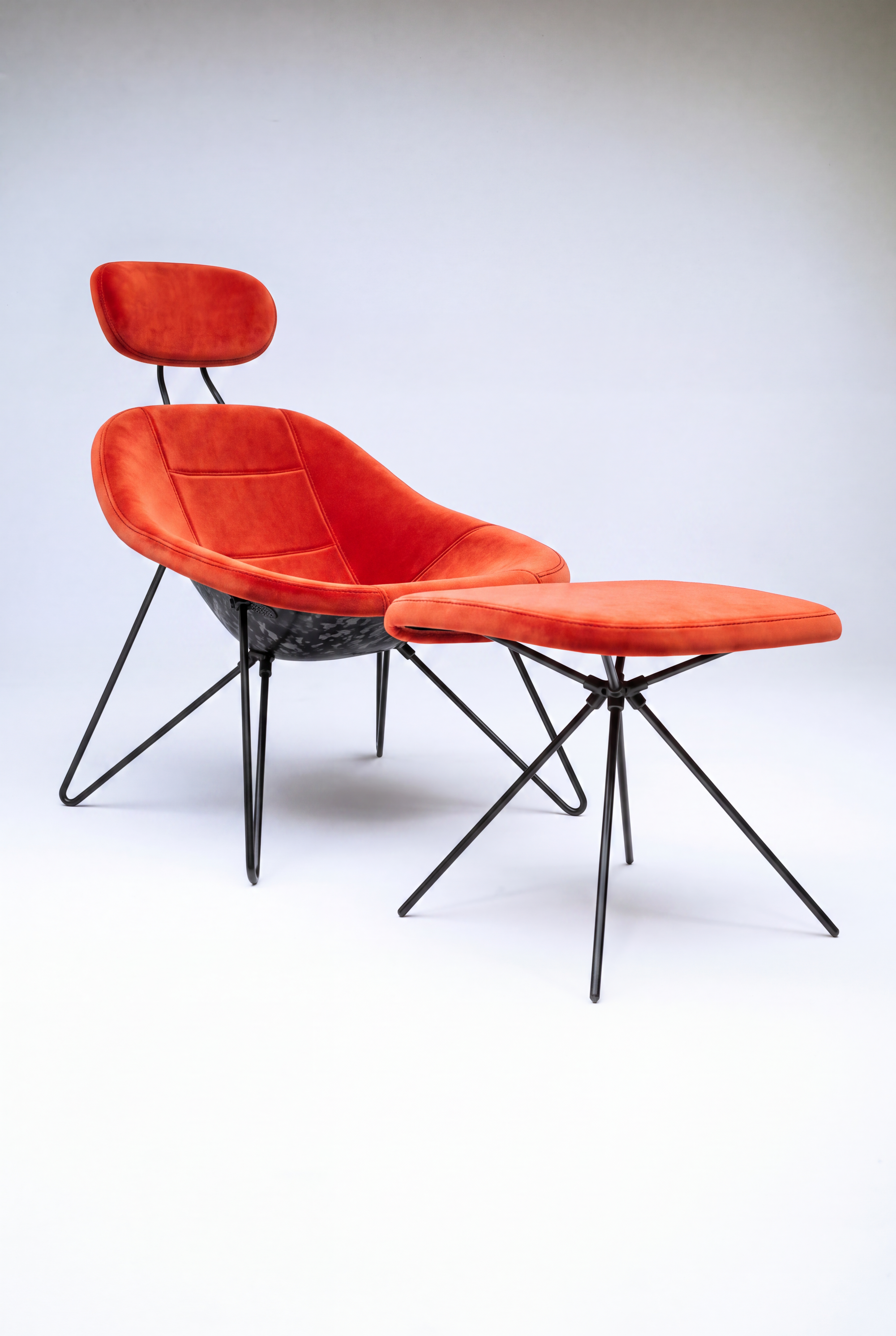House Dé Stains bespoke automotive furniture: Three-quarter view of the Maniac Lounge Chair and matching ottoman featuring bright orange-red velvet upholstery, a hand-finished carbon fiber composite shell, and matte black tubular steel frames.